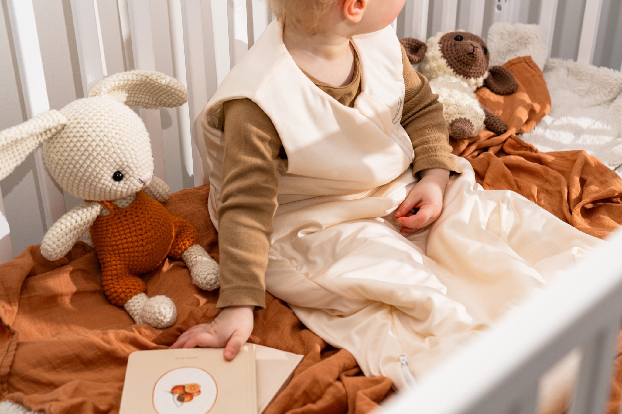 Toddler sitting in a crib wearing the Nömad sleep sack, surrounded by plush toys